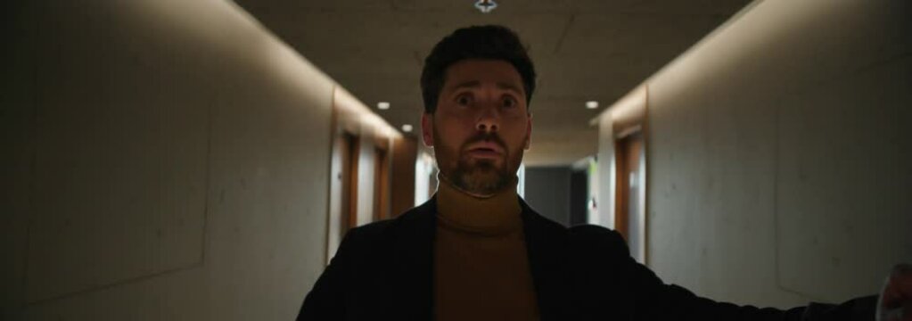 Portrait of young Caucasian man feeling dizzy while walking down hallway, holding onto wall. Businessman in formal clothes leaning on wall in hotel. Shot on camera with anamorphic lens