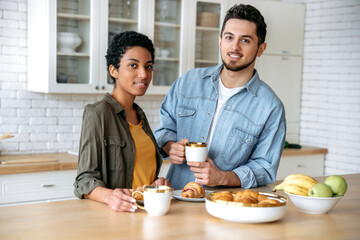 Fototapeta na wymiar Happy spouses of different nationalities, stand at home in the kitchen, dressed in stylish clothes, drinking morning coffee with croissants, discussing plans for the weekend, look at each other, smile