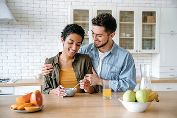 Happy married couple, caucasian man and african american woman, having breakfast at cozy home kitchen with healthy porridge, they happy to spend time together at weekend morning, communicating, smile