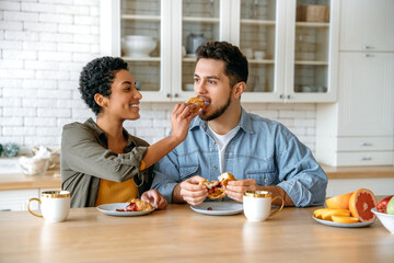 Fototapeta na wymiar Joyful couple in love of different nationalities, happy spouses, sit at home in the kitchen, drinking morning coffee with croissants, woman feeding a man a croissant, look at each other, smile