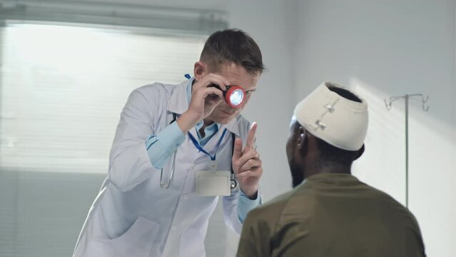 Doctor examining eyesight of soldier with bleeding head wound in military hospital with flashlight