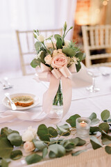 A beautiful floral bouquet in a vase with a bow on a table decorated with green garland at a wedding reception.