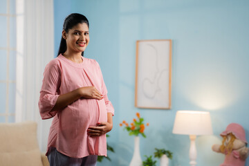 Happy smiling Indian Pregnant woman standing by feeling tummy while looking camera at home - concept of motherhood, expecting baby and parenthood