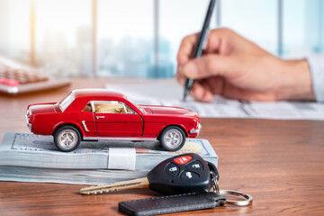 Car loan or buying a new car concept. Red car on dollar banknotes and car key on table with a man signing on paper in background. - obrazy, fototapety, plakaty