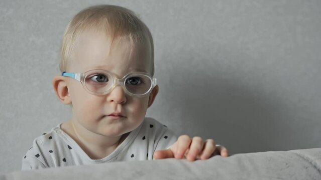 Serious one year old baby with squint and glasses is watching TV at home. Caucasian infant has strabismus poor eyesight or bad vision. White kid has an eye disease. Portrait of small child