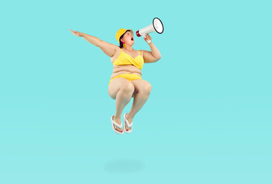 Funny Crazy Overweight Chubby Woman In Yellow Swim Suit, Sun Hat, Flip Flops With Fat Folds On Belly Jumps High Up In Air On Blue Background And Makes Loud Beach Holiday Announcement Through Megaphone