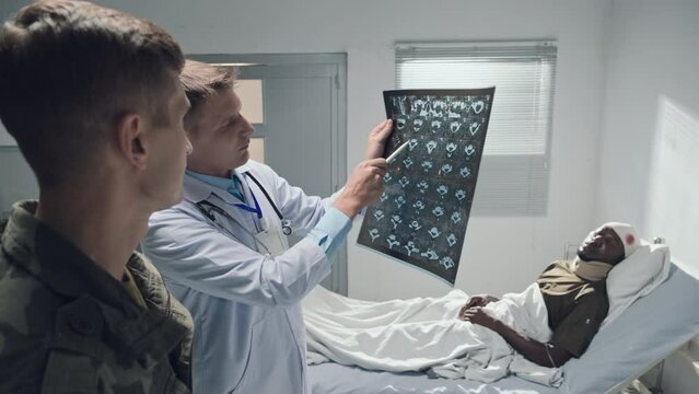 Doctor in white uniform explaining result of x-ray image of wounded patient to soldier in ward of military hospital