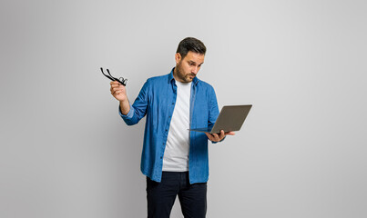 Shocked young businessman holding glasses and looking at laptop while standing on white background
