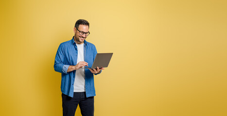 Smiling businessman wearing eyeglasses working on laptop while standing on yellow background - Powered by Adobe