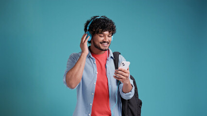 Hispanic or Arab young male student listening to music with headphones and smartphone on blue studio background