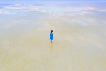 A woman in a blue dress walks along the shore of a salt lake with mirrored sky and clouds at sunset