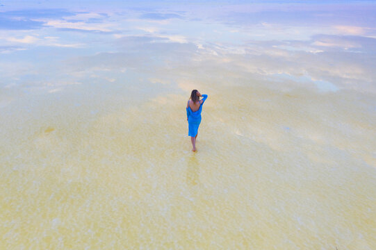A girl in a blue dress walks along the shore of a salt lake with mirrored sky and clouds at sunset