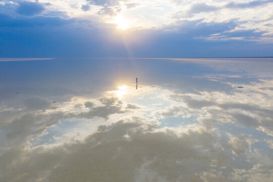 A woman in a blue dress walks along the shore of a salt lake with mirrored sky and clouds at sunset