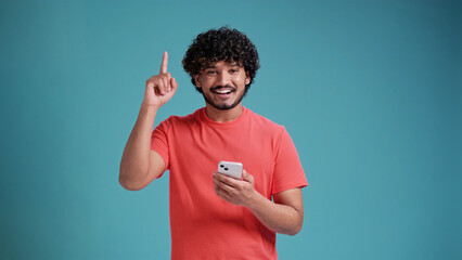 Young Hispanic or Arab man using smartphone standing very happy pointing with hand and finger showing up, in coral t-shirt on blue studio background