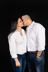 Bald man kissing his wife on the cheek as she smiles on a black background.