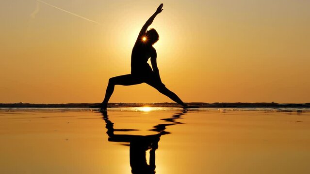 In picturesque setting of vibrant sunset sky, woman raises her hand and bends her body, performing Reverse Warrior yoga position. Silhouetted shot, against sunset sky on backdrop