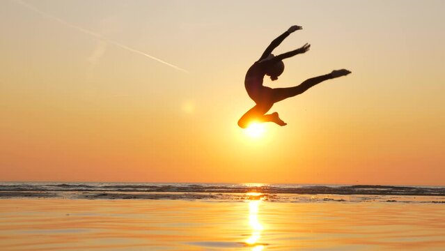 Young gymnast gracefully leaps into backbend, her silhouette contrasting against backdrop of stunning sea sunset. Skilled athlete performs captivating routine on sandy bank