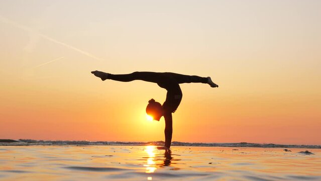 In picturesque setting of beautiful sunset beach, talented gymnast girl captivates with her impressive skills. With effortless grace, she performs handstand split, transitioning into single stag