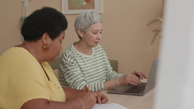 Waist up of two modern multiracial mature women using laptop and chatting sitting at desk in cozy bright peach walled room at daytime