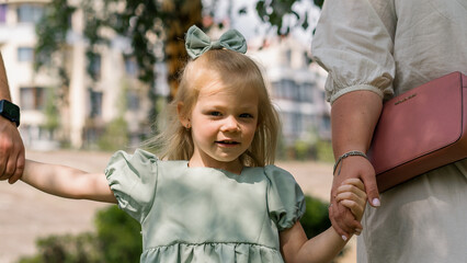 Close up portrait of cute little girl with bow walking in summer park holding hands of her parents Happy moments and family weekend