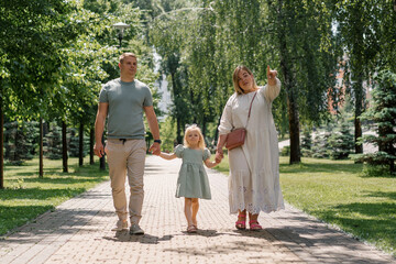 Naklejka na ściany i meble Little cute girl in green summer dress with parents in city park on a walk summer family lifestyle Childhood happy family holding hands