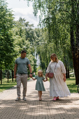 Little cute girl in green summer dress with parents in city park on a walk summer family lifestyle Childhood happy family holding hands