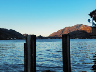 panorama of the waters of a lake, with 2 poles in the foreground and mountains in the background, a balcony with lanterns on the right, all at first light on a clear day