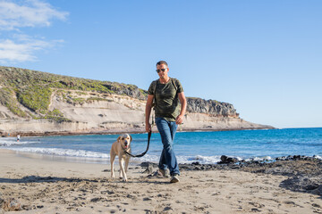 A traveling man walks the beach with his dog and wears a backpack