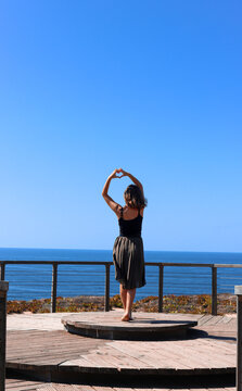 A female standing gracefully in a doll position with hand heart gesture. Graceful Ballerina Standing In Ballet Position in front of the ocean on a wooden platform.