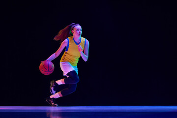 Young concentrated girl, basketball player in motion, running with ball against black studio...