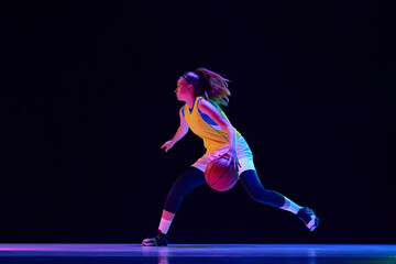 Young female athlete, professional basketball player in motion, dribbling ball over black studio background in neon light. Professional sport, action and motion, game, competition, hobby, ad concept