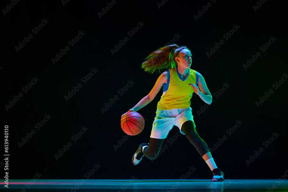 Wall mural Young motivated girl, basketball player in uniform running with ball against black studio background in neon light. Concept of professional sport, action and motion, game, competition, hobby, ad