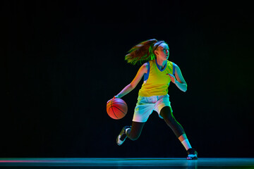 Young motivated girl, basketball player in uniform running with ball against black studio...