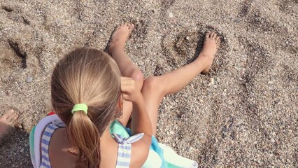 Little girl in stripy swimsuit sits on sandy beach and eats snacks after swimming, top down view