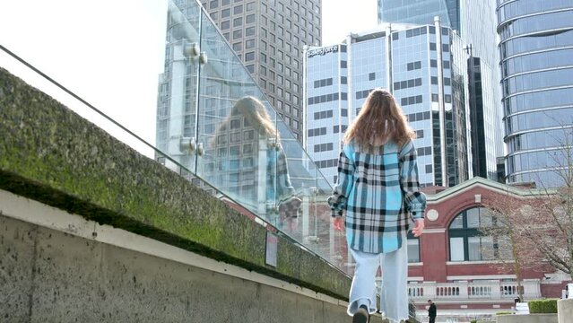 teenager girl walks along embankment against backdrop of skyscrapers of huge glass buildings back girl goes far stops and looks at nature Vancouver Canada Place Harbor Center