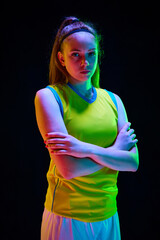 Portrait of young girl, basketball athlete posing in uniform with serious face against black...
