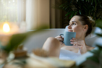 woman relaxing in bath and drink a coffee at home bathroom. looking out of window - 613954184