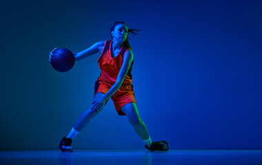 Dynamic image of young girl, basketball player in uniform in motion, playing over blue studio background in neon light. Concept of professional sport, action and motion, game, competition, hobby, ad