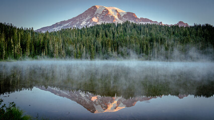 Mount Rainier and pine forest reflecting into Reflection Lakes at sunrise with light fog over lake | Mount Rainier National Park, Washington, USA