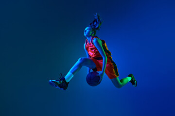 Obraz na płótnie Canvas Young sportive girl, basketball player jumping with ball against blue studio background in neon light. Concept of professional sport, action and motion, game, competition, hobby, ad
