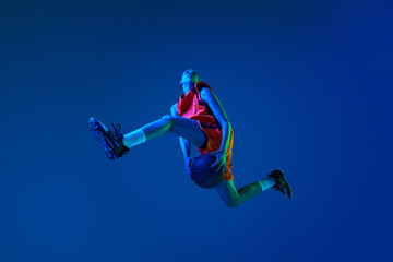 Bottom view of young girl, basketball player jumping with ball against blue studio background in...