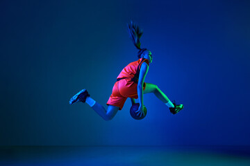 Fototapeta na wymiar Side view of young girl, basketball player jumping with ball against blue studio background in neon light. Concept of professional sport, action and motion, game, competition, hobby, ad
