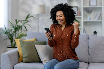 African american woman happy received online notification, good news , woman sitting at home on couch in living room and celebrating victory success, holding hand up gesture of triumph and achievement