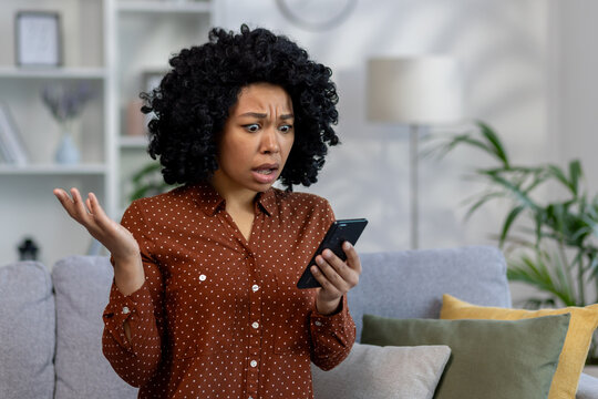 Sad woman alone at home, african american woman received notification message with bad news sitting on couch in living room, holding phone in hands using social media app
