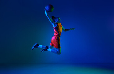 Slam dunk. Motivated young girl, basketball player throwing ball in jump against blue studio background in neon light. Concept of professional sport, action and motion, game, competition, hobby, ad