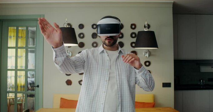 A man in casual clothes puts on a virtual reality headset and starts moving his hands while standing in a stylish living room