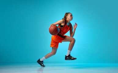 Fototapeta na wymiar Young athletic girl, basketball player in motion, running with ball against blue studio background in neon light. Concept of professional sport, action and motion, game, competition, hobby, ad