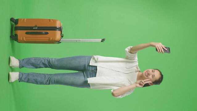 Full Body Of Asian Female Traveler With Luggage Smiling And Taking Photo On Smartphone While Standing In The Green Screen Background Studio
