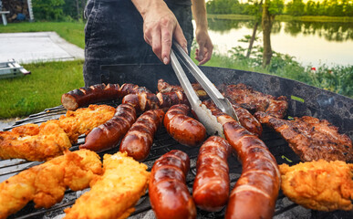 Man turning the sausage on the bbq with kitchen tongs. Person grilling in the garden. Barbeque with sausages and fried fish on the grill. 