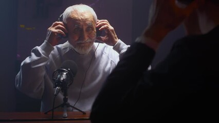 The old man creates a podcast in a professional studio. Senior male does online streaming interviews. Elderly man blogger speaking in microphone or make vlog. Recording audio content for social media